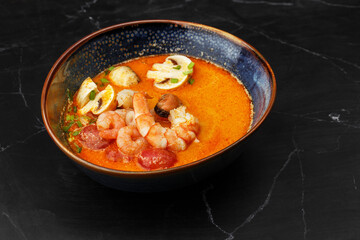 Asian soup Tom Yam with shrimps, mussels, mushrooms, tomatoes and spring onion. Dish isolated in a blue bowl, close-up on a black marble background. Asian cuisine.