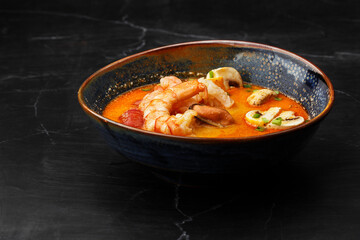 Asian soup Tom Yam with shrimps, mussels, mushrooms, tomatoes and spring onion. Dish isolated in a blue bowl, close-up on a black marble background. Asian cuisine.