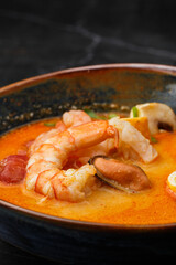 Asian soup Tom Yam with shrimps, mussels, mushrooms, tomatoes and spring onion. Dish isolated in a blue bowl, close-up on a black marble background. Asian cuisine.
