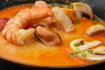 Asian soup Tom Yam with shrimps, mussels, mushrooms, tomatoes and spring onion. Dish isolated in a blue bowl, close-up on a black marble background. Asian cuisine.