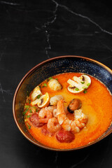 Asian soup Tom Yam with shrimps, mussels, mushrooms, tomatoes and spring onion. Dish isolated in a blue bowl, close-up on a black marble background. Asian cuisine.