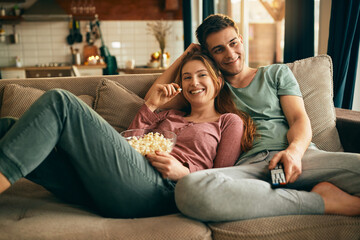 Happy couple eating popcorn and watching TV while relaxing on the sofa. - obrazy, fototapety, plakaty