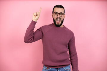 Handsome man wearing glasses and casual clothes over pink background pointing finger up with...