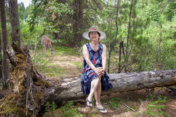 lady sitting on a log resting with a deer in the background in the forest.