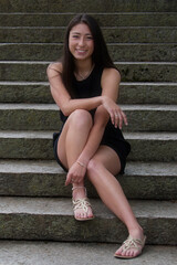 Full body outdoor portrait of beautiful young woman in sleeveless black dress smiling and looking towards the camera while sitting on cement stairs