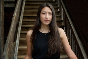 Outdoor portrait of beautiful young woman in sleeveless black dress smiling and standing at the bottom of wooden staircase.