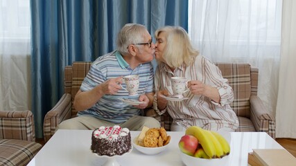 Elderly couple holding drinking tea talking, laughing, smiling, kissing, sitting on sofa at home