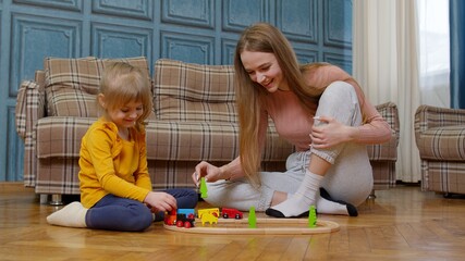 Mother with little daughter child girl riding toy train on wooden railway blocks board game at home