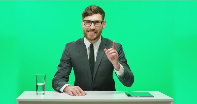 Successful male TV presenter sitting in studio at table with tablet and glasses of water on background of chromakey telling breaking news.