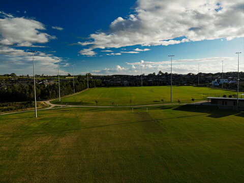 Aerial photograph of suburban sporting field and housing estate at sunset, with blue sky and white clouds.