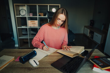Cute student studies online at home, rewrites information from a book in a notebook with a serious face. Female student doing homework at home.
