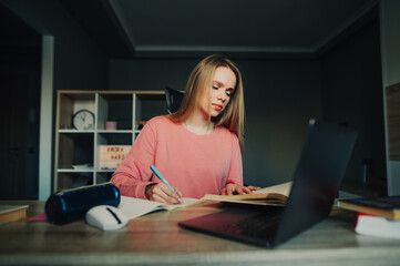 Beautiful female student sitting at a desk at home near a laptop and rewrites a pen from a book in a notebook with a serious face. Concentrated woman studying at home.