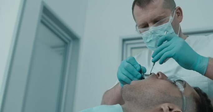 In dental office a male patient laying in the dental chair with open mouth while the male dentist in disposable mask and glasses holding inside of the mouth a male patient a dental impression.