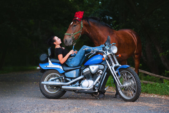 The young dark-haired woman lies on a blue motorcycle and holds a number of his horse. Horsewoman with the living and the iron horses.The horse at the head bandana and motorcycle goggles.