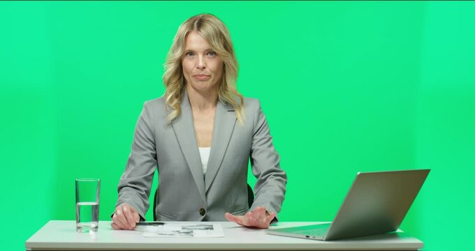 Popular female TV presenter in gray suit sitting in studio at table with laptop glasses of water on background of chromakey and telling good news.