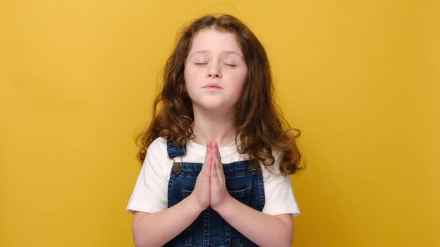 Hopeful cute little girl kid keep hands in prayer hope believe ask good luck, smiling small child believer pray do religious superstitious gesture, isolated on yellow studio background. Hope concept