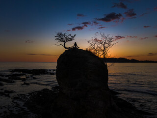 Beautiful and peaceful aerial view of a woman practicing yoga and meditation on a rock over the sunset in a beach
