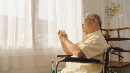 Asian male patient looking outside the window while sitting in wheelchair in living room at retirement home. Asian Thai elderly person sitting lonely in a wheelchair holding a paper bird to play.