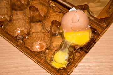Transparent plastic egg tray with one broken brown egg on table. Half egg with yolk in an empty box. Yolk overflowed into the adjacent cavity. Сracked eggshell.