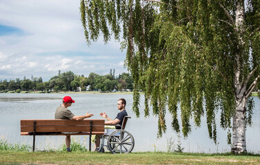 caucasian father and grown son in a wheelchair sitting by a lake.