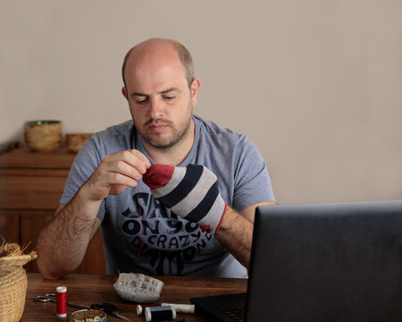 Emancipated man darning his socks in the kitchen of his home. On the table are a laptop computer and sewing supplies. Copy space.