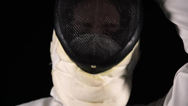 Portrait of a confident fencer looking at the camera and putting on a protective mask on a black studio background. Female athlete in a white uniform posing with a rapier in her hands. Close up.