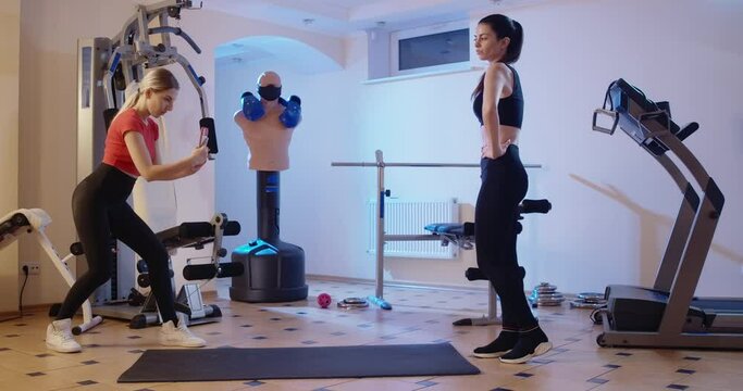 Wide shot of two confident slim millennials taking photos in gym indoors. Gorgeous slender Caucasian women posing after training workout. Femininity and modern lifestyle. Cinema 4k ProRes HQ.