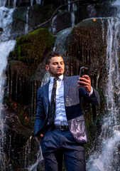 Fototapeta premium Telework employee concept. Handscome business man in suit holding smartphone telephone mobile phone at waterfall background.