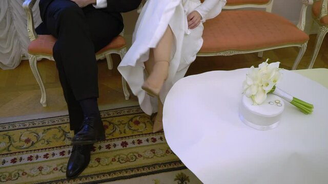 Bride and groom sitting on sofa indoors and crossing legs. Wedding bouquet of flowers on background. High quality FullHD footage