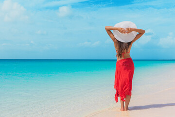 Beach summer vacation free relaxing woman enjoying sun wearing hat and red skirt with arms behind standing at the blue idyllic ocean. Paradise getaway holiday. - obrazy, fototapety, plakaty