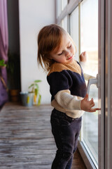 A little girl stands on the windowsill. The child holds the window opening handle.