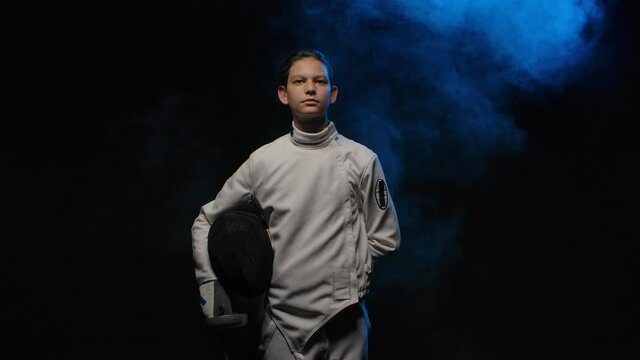 Portrait of young guy swordsman putting on protective mask helmet. Athlete in white uniform posing with rapier in hand against smoky black studio background with blue light. Slow motion. Close up.