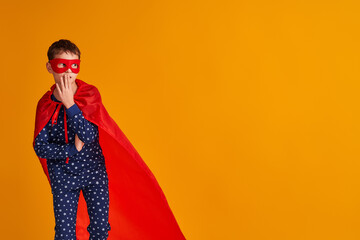 surprised child is playing in a superhero costume. the boy is dressed in a red cloak and mask, holding his hand to his mouth and looking doubtfully to the side on a yellow background. Copy space