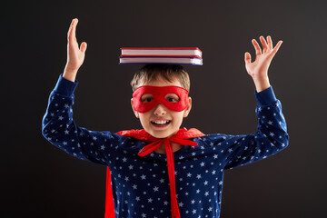 cheerful child dressed in a superhero costume, holding books on his head, a little boy laughs and enjoys studying at school. Posing on a black background in the studio. Forward to knowledge. Close-up
