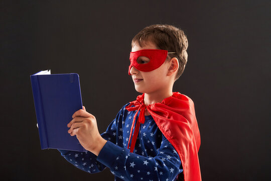 attentive child, dressed in superhero costume, holds books in hand and reads enthusiastically. boy is interested in new book, he is happy to get knowledge. Learn to read quickly on black background.