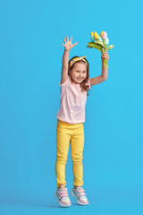 charming little girl with curly hair and headband with bouquet colorful tulips made fabric jumps to her full height on blue background studio. child in fashionable summer clothes. Dynamic image