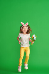 charming happy girl with Easter bunny ears jumps on a green background in full growth. a child smiles and holds a bouquet of fabric tulip flowers on Easter Day. The joy of the arrival of spring.