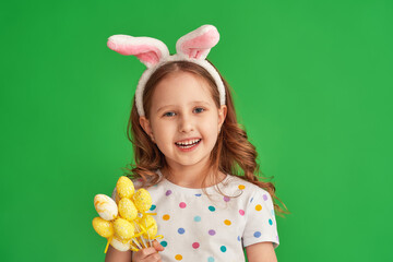 Portrait of a cute little girl wearing the ears of an Easter bunny holding colorful eggs standing on a green background on Easter day. The child is happy and celebrates Easter