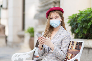 Asian woman wearing suit and red cap holds smartphone in her hands while sits on bench in park...