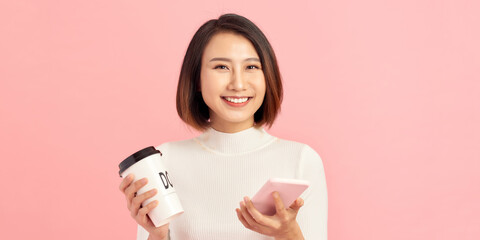 Young asian woman over isolated pink background holding coffee to take away and a mobile