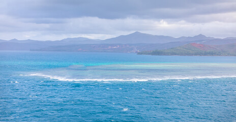 Beautiful Turquoise water surrounding a small coral reef close to the shore of New Caledonia with mountains in the background covered in haze and clouds.