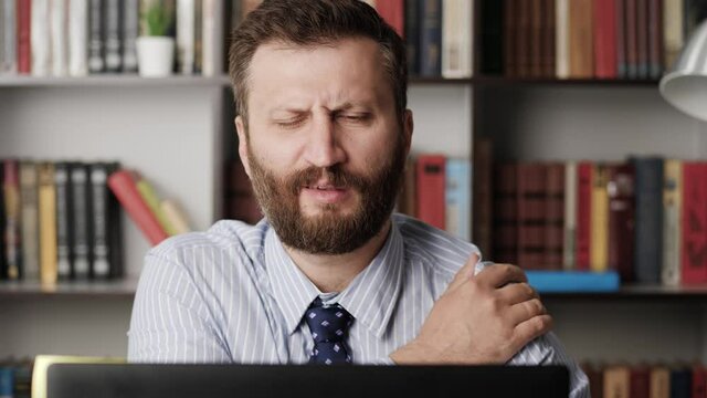 Shoulder pain. Bearded man working at computer and suddenly feels pain and discomfort in shoulder, he touches his shoulder with hand and stretches. Slow motion