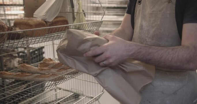 Male baker putting freshly baked sourdough bread in paper bag for customer in bakery shop