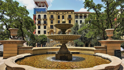 Obraz premium Fountain in the city square. Water splashes down the stone bowls. Round pool. Nearby are green trees, a residential building. South Africa. Johannesburg