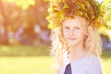 Fototapeta premium Little girl with autumn maple wreath. Yellow and green colors. Backyard nature. Female child portrait. Happy people. Model posing at park
