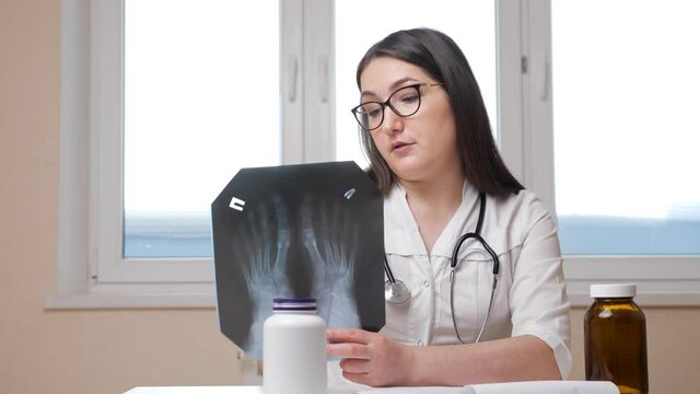 Woman doctor with stylish glasses and stethoscope shows X-ray picture of strange feet to camera sitting at table in hospital office closeup