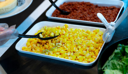 Selective focus on sweet corn in white bowl at salad bar buffet of restaurant. Hand wear plastic gloves holding black spoon scoop yellow sweet corn. Healthy eating. Healthy food. Vegetarian food.