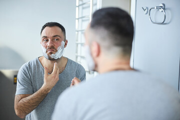 Obraz premium Bearded man applying shaving cream on face in bathroom