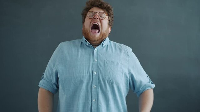 Portrait of guy in glasses sneezing looking at camera standing against gray background alone wearing casual clothing. Disease symptoms and bad health concept.