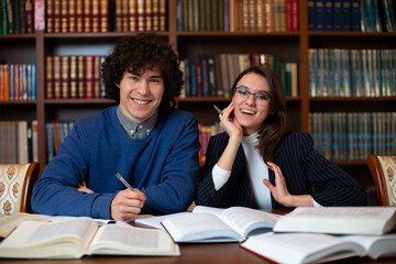 Two cheerful students perform together the project sitting in the library against the backdrop of bookcases. Student science. High quality photo